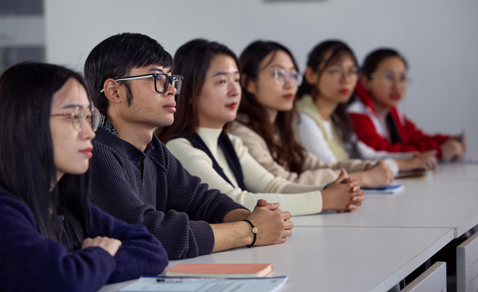 Six personnes assises à une longue rangée de bureaux, le regard tourné vers l’avant de la classe
